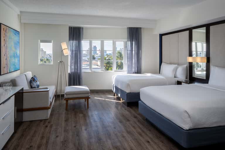 Bright hotel room with two queen beds, lounge seating, desk area, and expansive windows overlooking cityscape