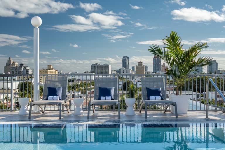 Rooftop pool deck with loungers, tropical plants, and panoramic city skyline views on clear blue day