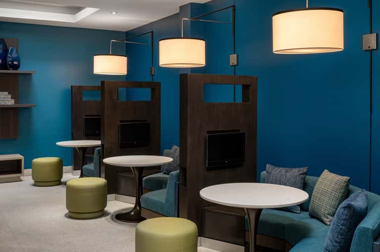 Hotel lounge with teal walls, wooden booth seating, cream pendant lights, lime poufs, and casual gathering spaces
