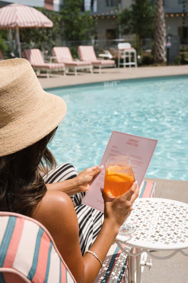 Guest holding pink menu and tropical drink poolside wearing straw hat