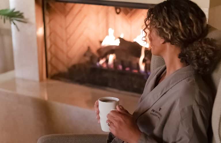 Woman in spa robe holding coffee cup, gazing at fireplace in resort setting