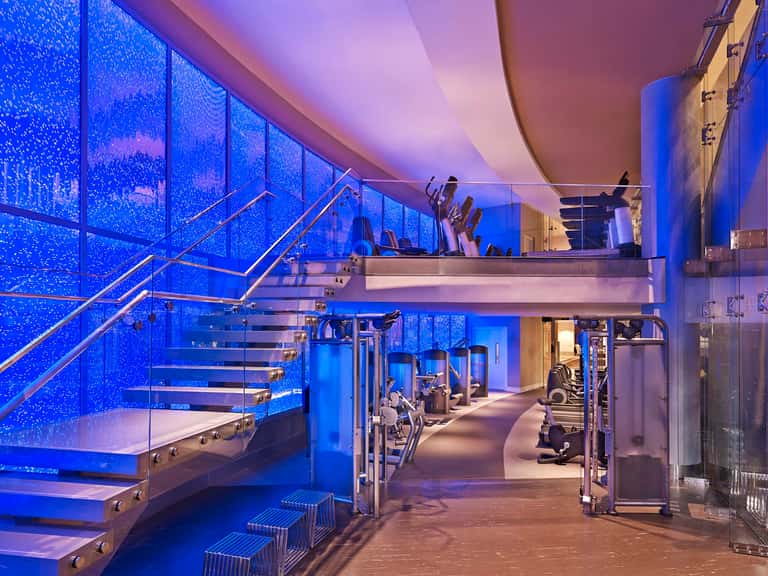 Modern gym with blue-lit windows, staircase, and exercise equipment on multi-level floors