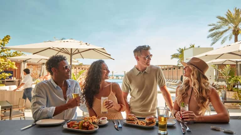 Four guests dining and socializing on a beachfront terrace with umbrellas and ocean views