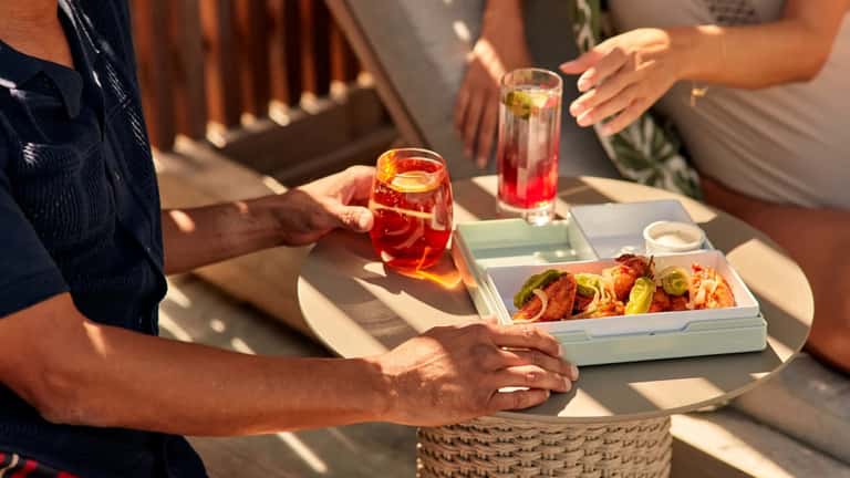 Close-up of hands holding tropical drinks and appetizers on a sunny resort terrace