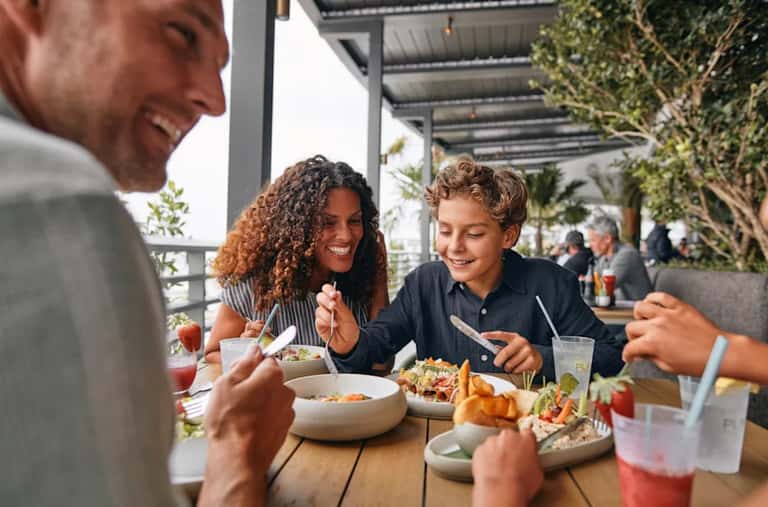 Family enjoying casual meal together on a resort patio with tropical plants and ocean views