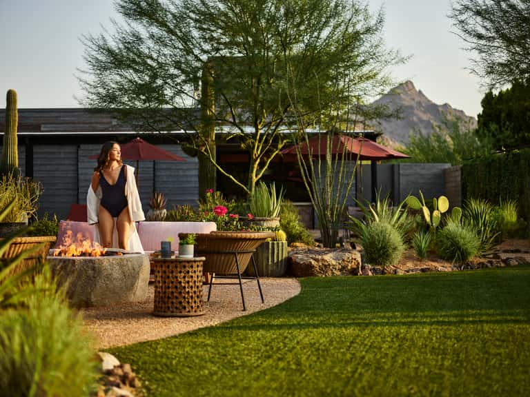 Woman in swimwear by fire pit at dusk with modern desert resort buildings and mountain backdrop