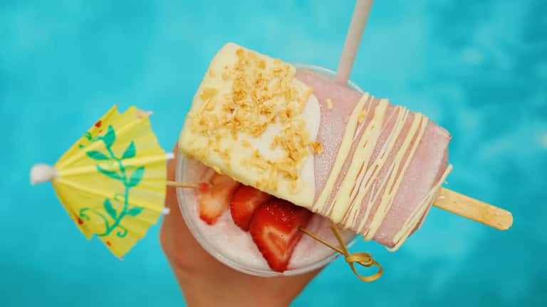 Colorful frozen popsicle with pineapple and strawberry garnish held over tropical drink