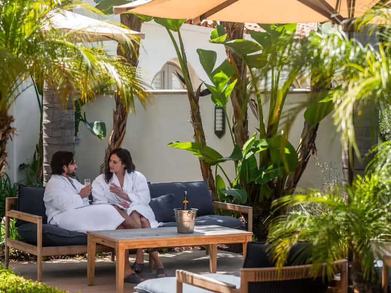 Outdoor spa lounge with couple on dark sofa surrounded by tall tropical plants and tan umbrellas