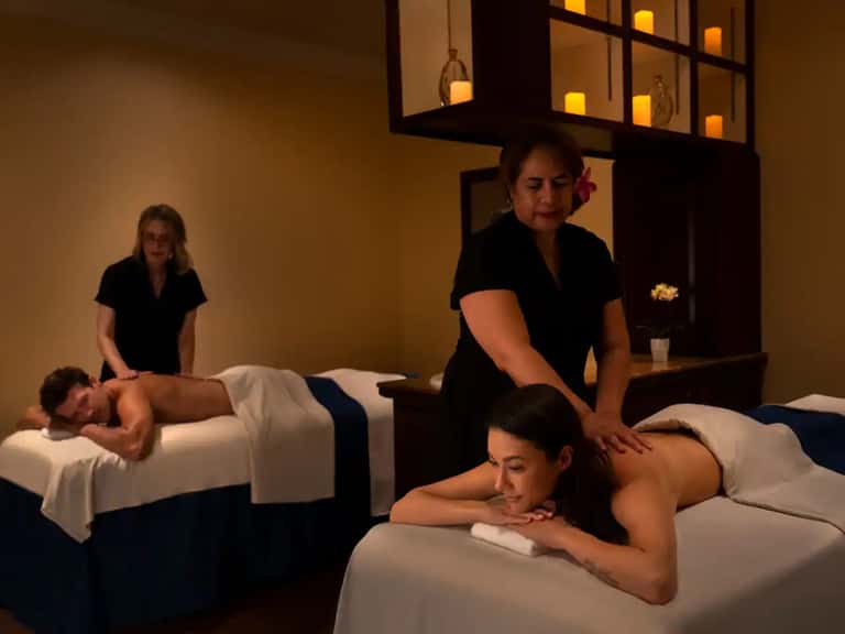 Spa treatment room with therapists providing massages to guests on white massage tables with warm lighting