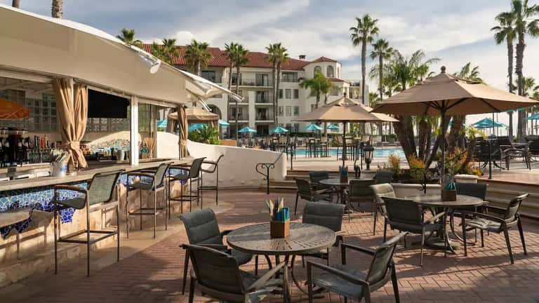 Poolside bar and lounge area with umbrellas, cabanas, palm trees, and resort buildings in background