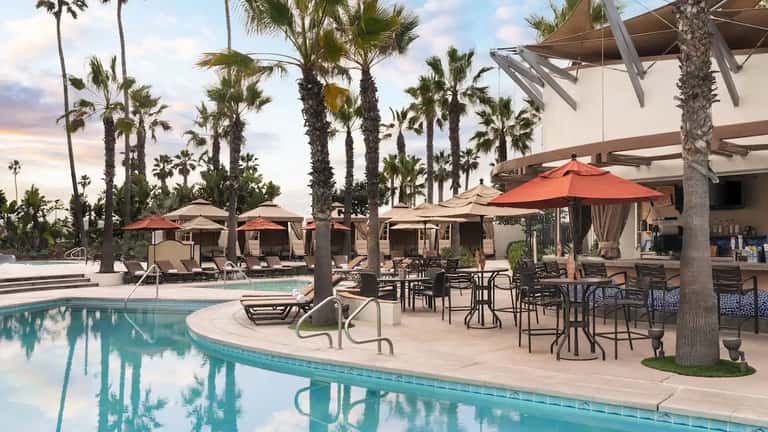 Resort pool deck with lounge chairs, umbrellas, palm trees, and modern shade structures at sunset