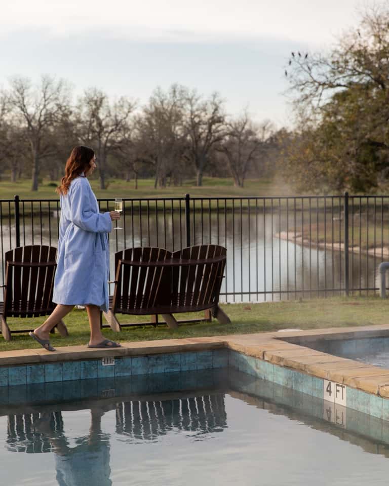 Woman in blue shirt holding wine glass standing by spa pool, with black Adirondack chairs and river view