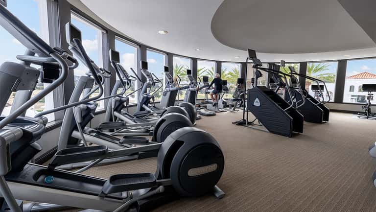 Modern fitness center with rows of elliptical machines and exercise equipment, floor-to-ceiling windows overlooking grounds