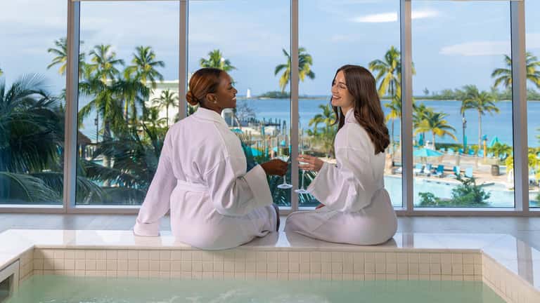 Two women sitting by indoor hot tub wearing white robes overlooking beach and palm trees