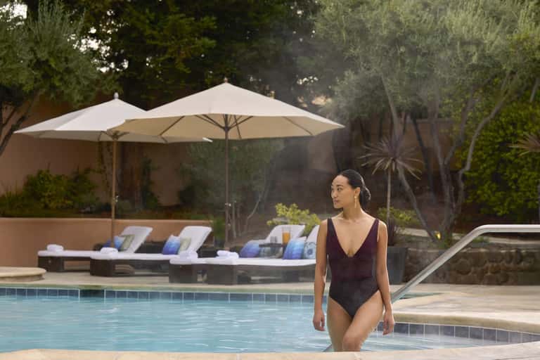 Woman in swimsuit entering outdoor resort pool with lounge chairs and umbrella in background