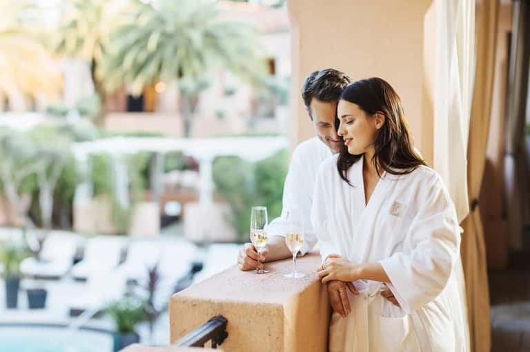 Couple in white robes enjoying champagne on terrace overlooking resort courtyard and gardens