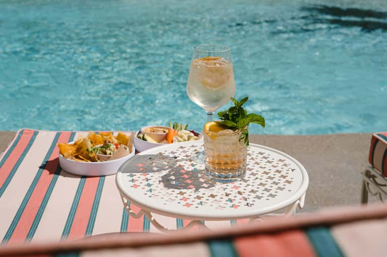 Poolside table with cocktail, appetizers, and pink menu on turquoise water background