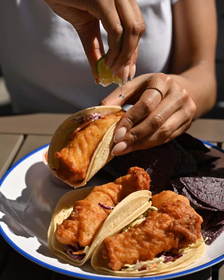 Hands assembling fried fish tacos with purple cabbage slaw on corn tortillas with lime wedge