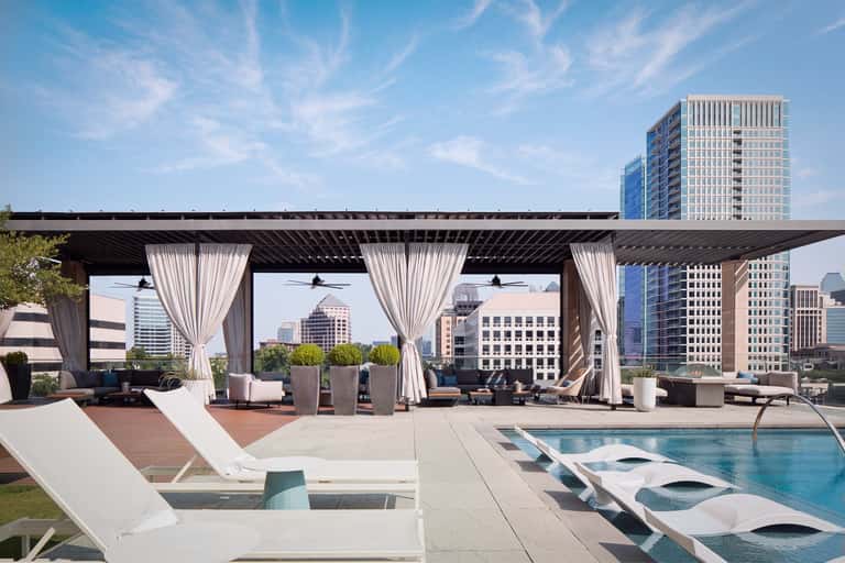 Rooftop lounge with pergola, curtains, lounge seating, infinity pool, and downtown skyline at daytime