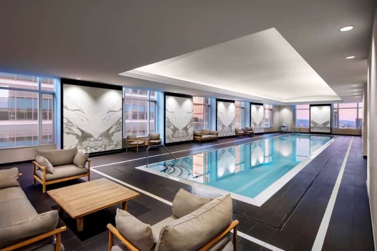 Luxurious indoor swimming pool with floor-to-ceiling windows, marble accents, lounge seating, and city skyline views