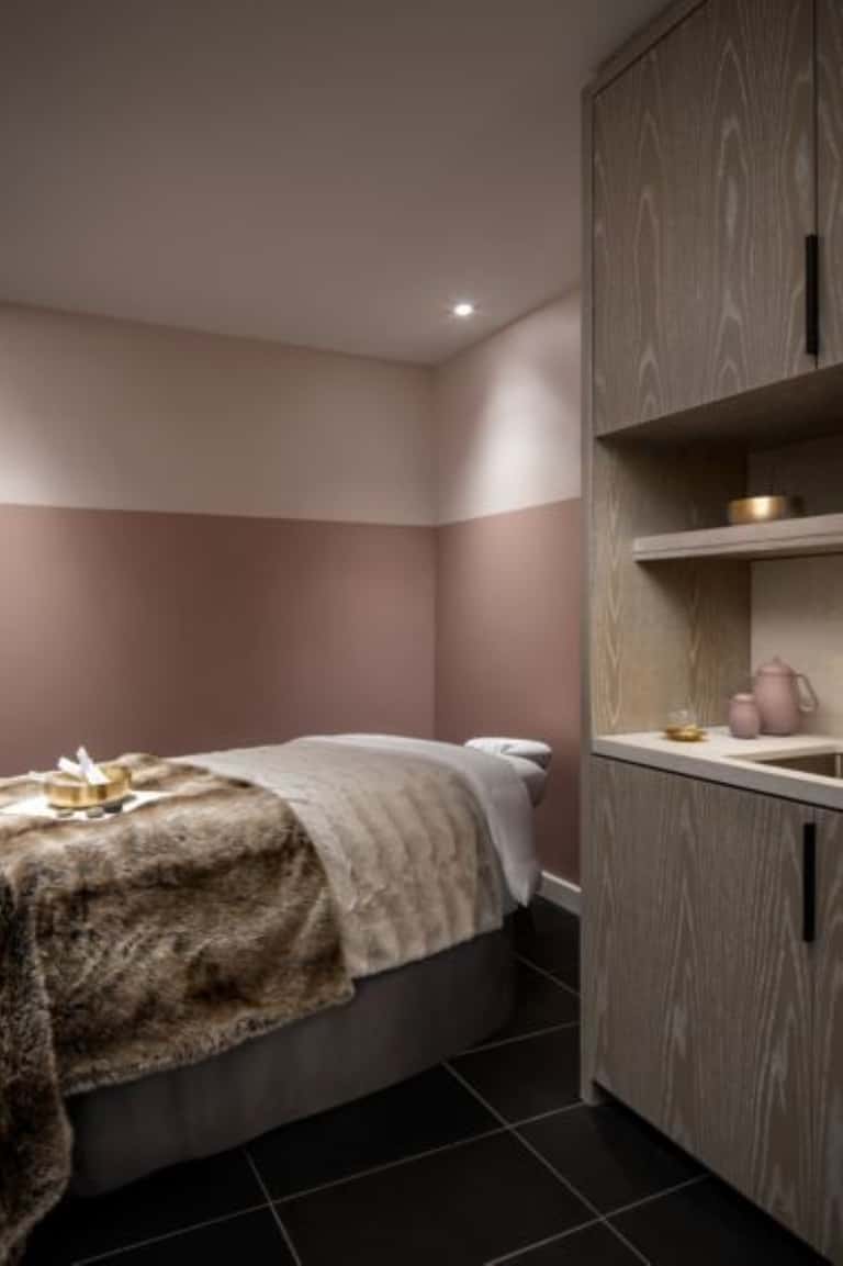Compact hotel bedroom with pink accent wall, wood cabinetry, bed, and integrated shelving
