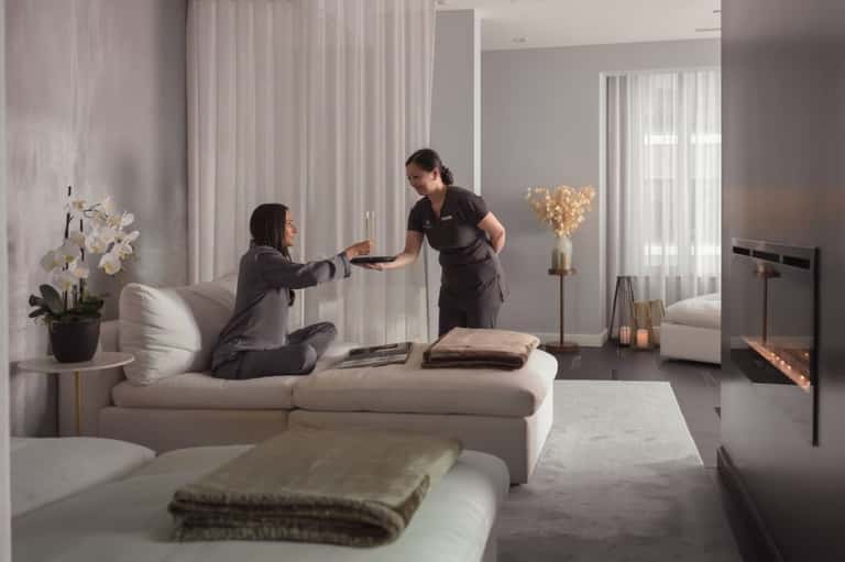 Spa attendant presenting towels to guest in modern wellness room with neutral tones and fireplace