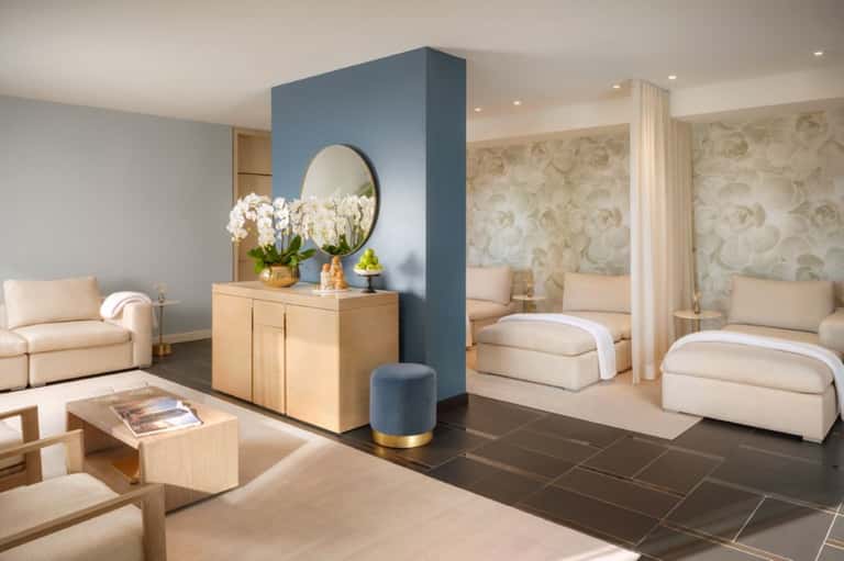 Luxury spa lounge with cream seating, blue accent wall, wooden dresser with white orchids, and patterned wallpaper