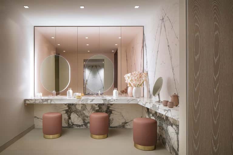 Luxury spa bathroom with marble vanity, dual illuminated mirrors, rose-toned stools, and wood-grain accent wall
