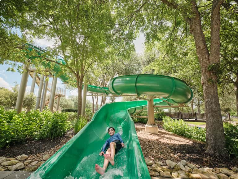 Child sliding down green water slide in wooded park setting with trees and playground equipment