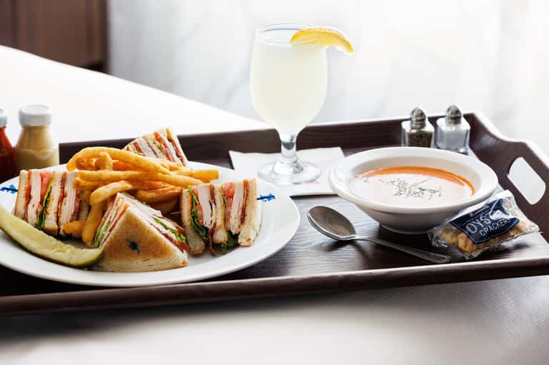 Hotel room service tray with club sandwich, french fries, soup, and lemon water on dark wood table
