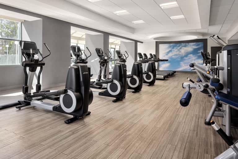 Bright fitness facility featuring rows of stationary bikes, cardio machines, and large screen display