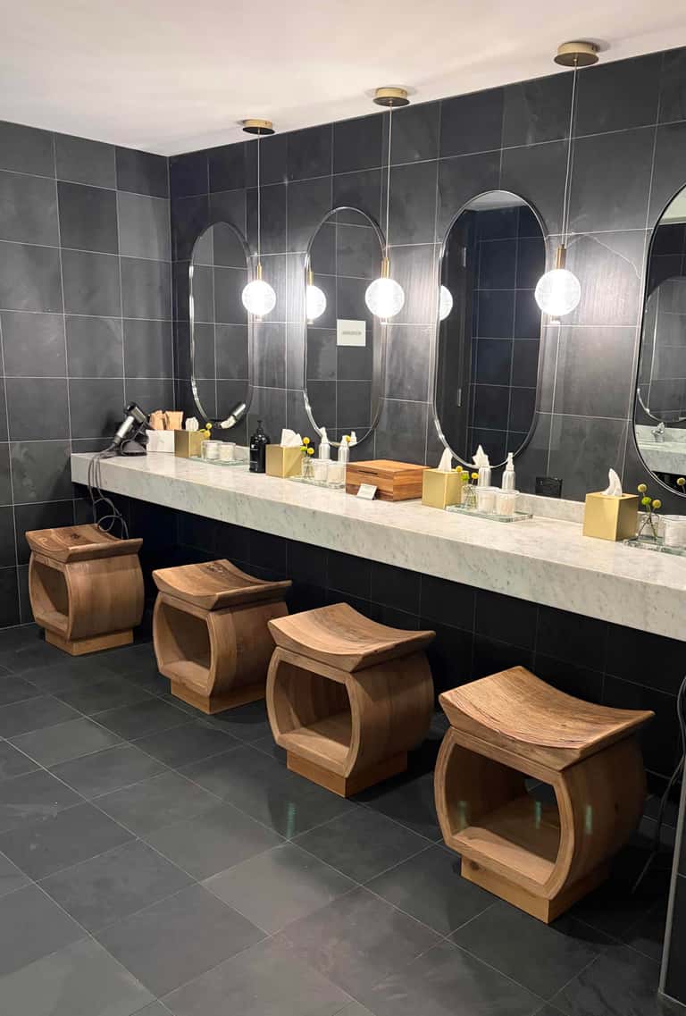 Spa bathroom with dark tiled walls, marble countertop, oval mirrors, and wooden stools