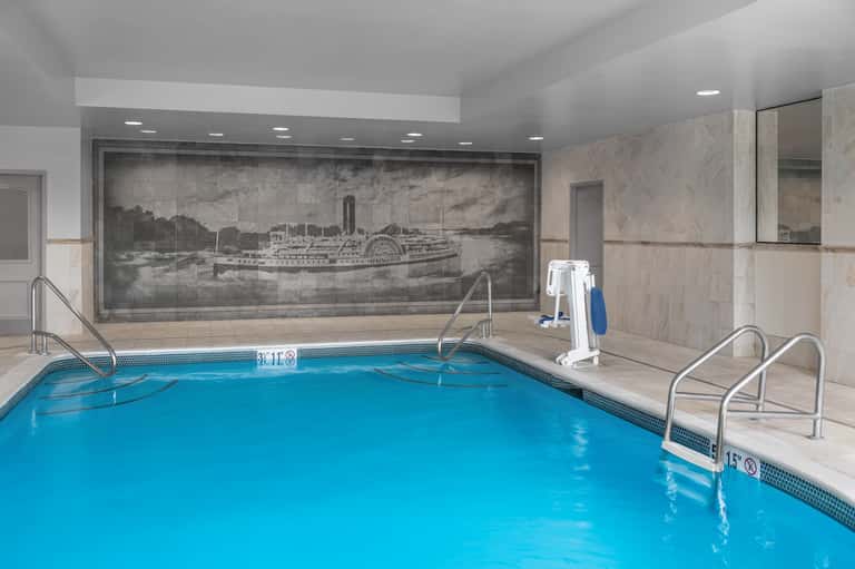 Indoor swimming pool with vintage steamboat mural, beige tile, and accessible railings