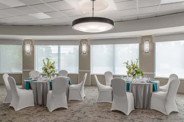 Banquet room with round tables, white chair covers, teal napkins, and large windows
