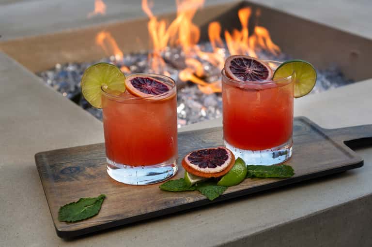 Two blood orange cocktails with citrus slices and mint on wooden board with fireplace background