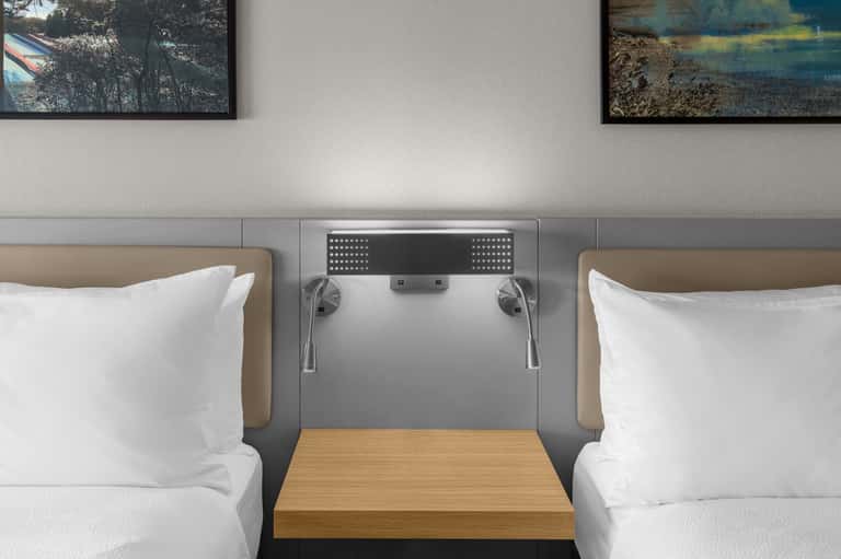 Two adjacent queen hotel beds with tan headboards and landscape artwork above