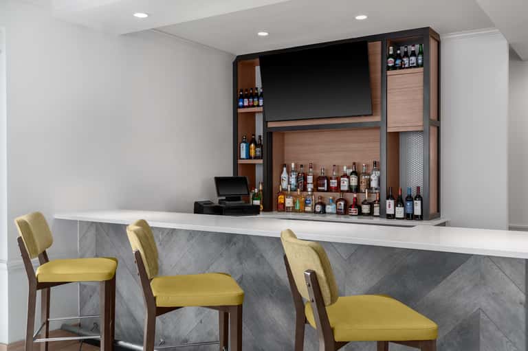Modern hotel bar with wooden shelving, premium liquor collection, yellow bar stools, and flat-screen TV