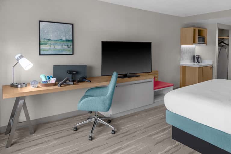 Hotel room workspace with desk, turquoise chair, TV, and queen bed with teal bedding