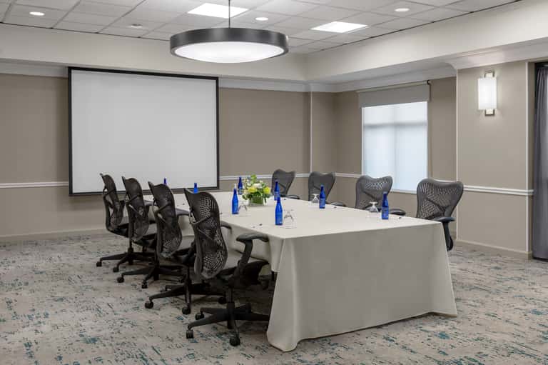 Conference room with long rectangular table, black mesh chairs, projection screen, and modern lighting