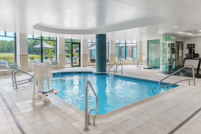 Indoor swimming pool with blue water, columns, lounge chairs, and large windows overlooking grounds