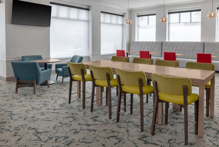 Meeting room with long wooden table, yellow chairs, gray banquette seating, mounted TV, and natural light from windows.