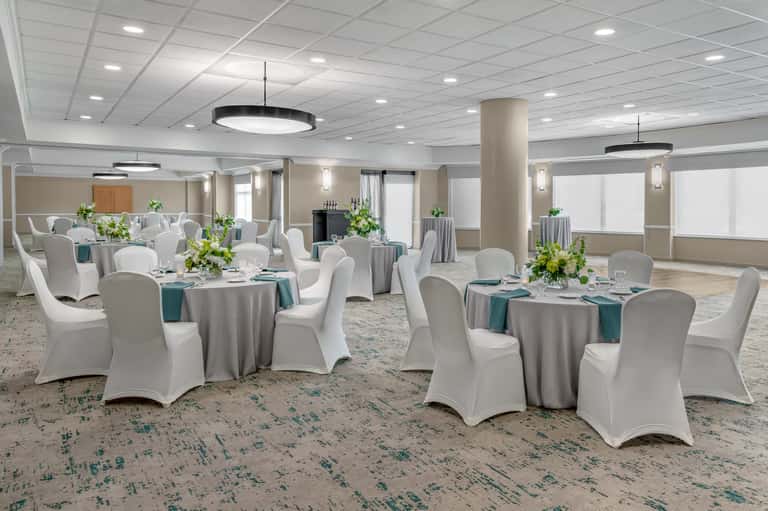 Ballroom setup with multiple round tables, white chair covers, teal linens, and overhead pendant lighting.