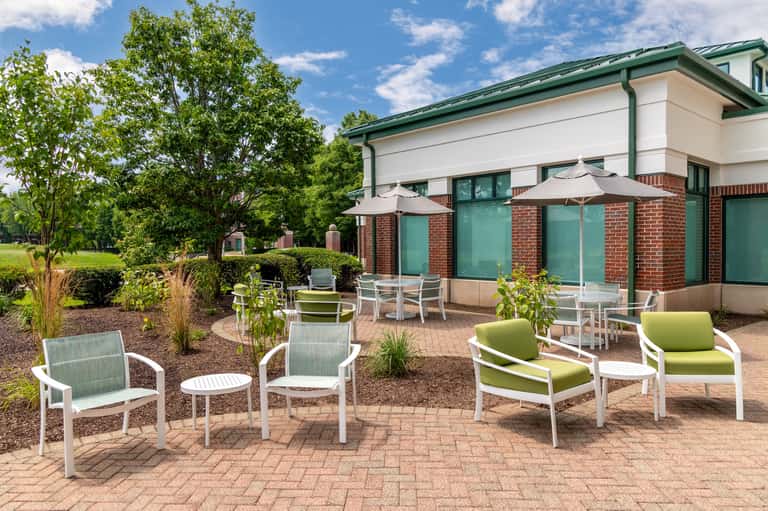 Patio seating area with white and green chairs, round tables, and brick building with green awnings.