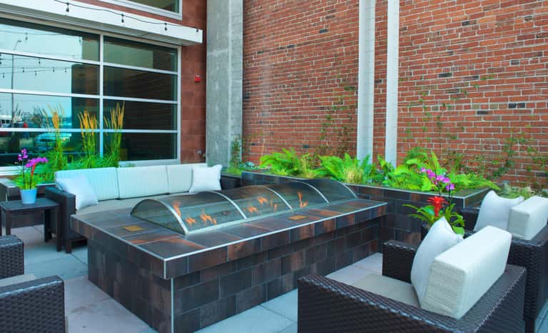 Modern outdoor patio with dark fire pit, white cushioned lounge chairs, and brick building backdrop with purple flowers