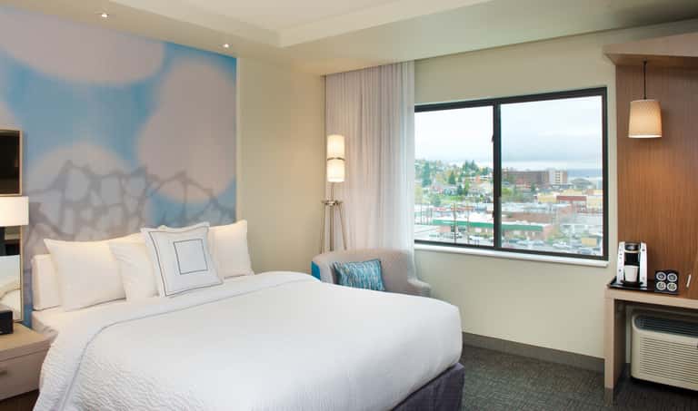 Hotel bedroom with queen bed, blue abstract wall art, city skyline visible through large windows, modern furnishings