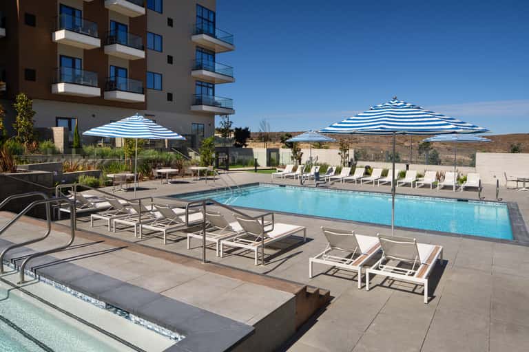 Resort pool with striped umbrellas, white lounge chairs, and desert hillside views at daytime