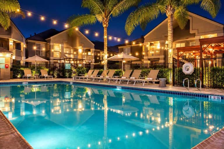 Illuminated resort pool at night with lounge chairs, palm trees, string lights, and residential buildings