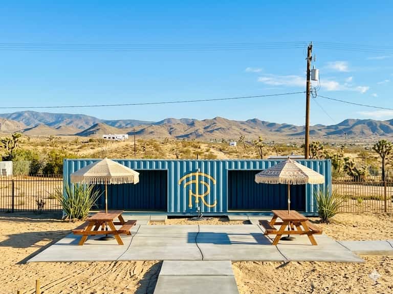 Modern shipping container structure with blue corrugated metal and gold logo in desert landscape