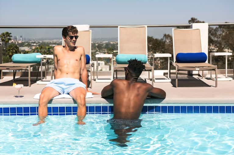 Two men relaxing in shallow resort pool with lounge chairs and city skyline views in background