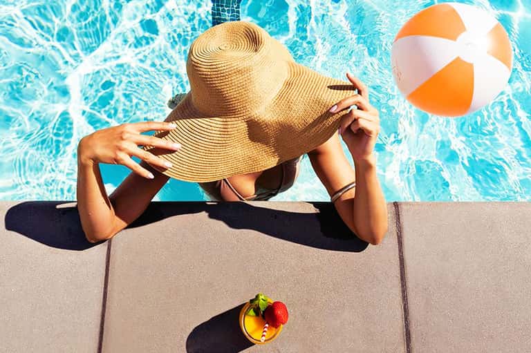 Woman in straw hat lounging poolside with beach ball and tropical drink